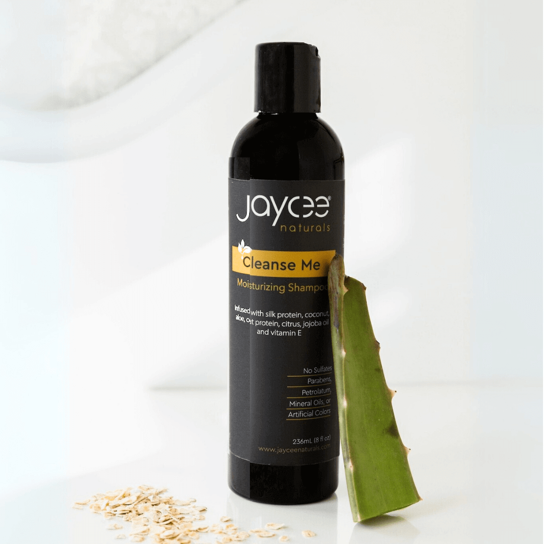 Jaycee Naturals Cleanse Me Moisturizing Shampoo, infused with silk protein and aloe and free of sulfates, parabens, petrolatum, mineral oils, and artificial colors. 236ml bottle standing on a white countertop and surrounded by oats and an aloe vera leaf.