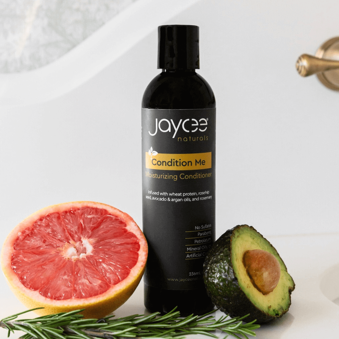 Jaycee Naturals Condition Me Moisturizing Conditioner, infused with wheat protein and argan oils and free of sulfates, parabens, petrolatum, mineral oils, and artificial colors. 236ml bottle standing on a white countertop surrounded by avocado and grapefruit.
