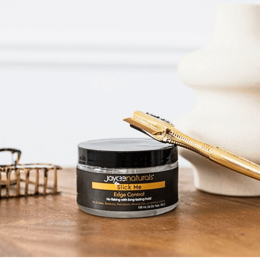 Jaycee Naturals Slick Me Edge Control, free of flaking and with a long-landing hold, and free of sulfates, parabens, petrolatum, mineral oils, and artificial colors. 118ml jar standing on a white countertop surrounded by a hair styling tool and hair accessory.