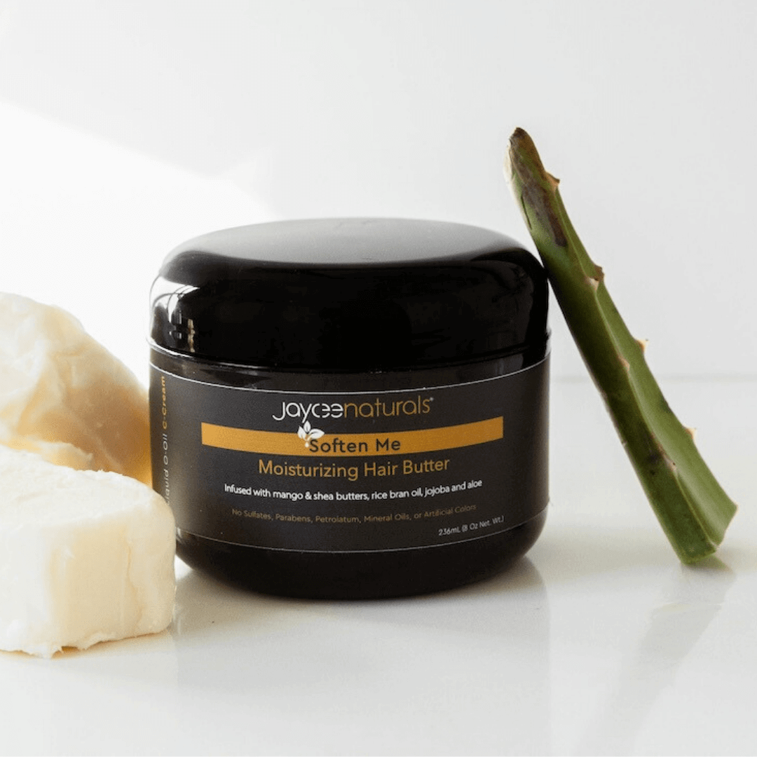 Jaycee Naturals Soften Me Hair Butter, infused with mango and shea butters and free of sulfates, parabens, petrolatum, mineral oils, and artificial colors. 118ml jar standing on a white countertop surrounded by shea butter and an aloe vera leaf.