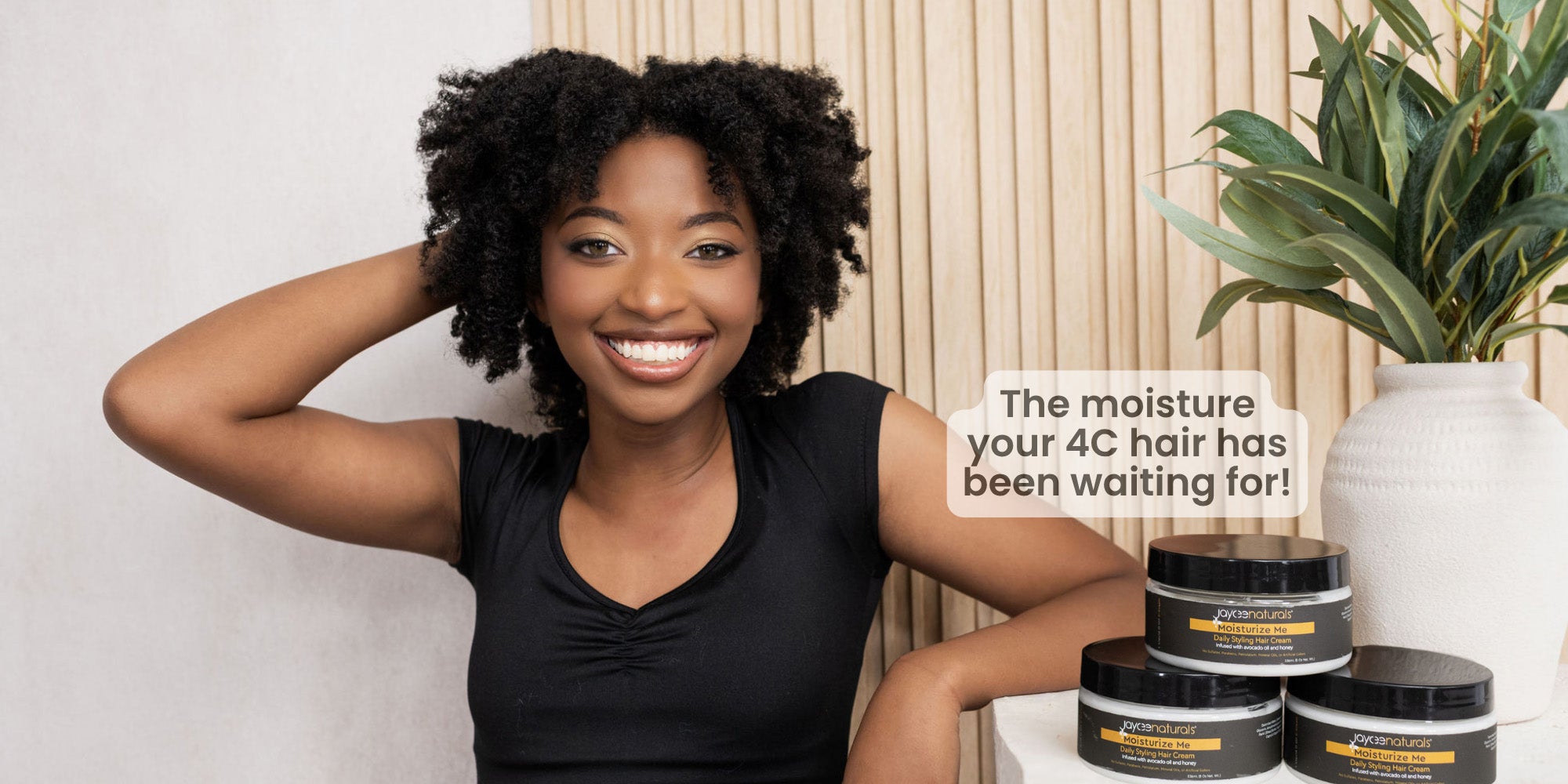 Woman with styled hair sitting next to hair care products with a plant in the background