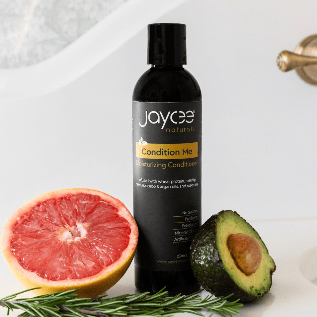 Jaycee Naturals Condition Me Moisturizing Conditioner, infused with wheat protein and argan oils and free of sulfates, parabens, petrolatum, mineral oils, and artificial colors. 236ml bottle standing on a white countertop surrounded by avocado and grapefruit.