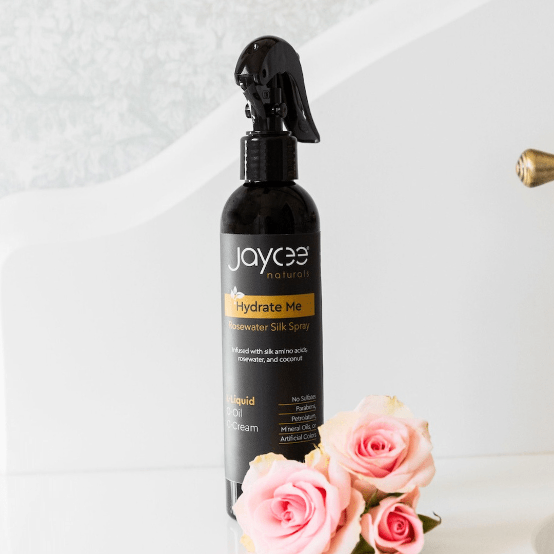 Jaycee Naturals Hydrate Me Rosewater Silk Spray, infused with silk amino acids and free of sulfates, parabens, petrolatum, mineral oils, and artificial colors. 236ml bottle standing on a white countertop surrounded by pink roses.