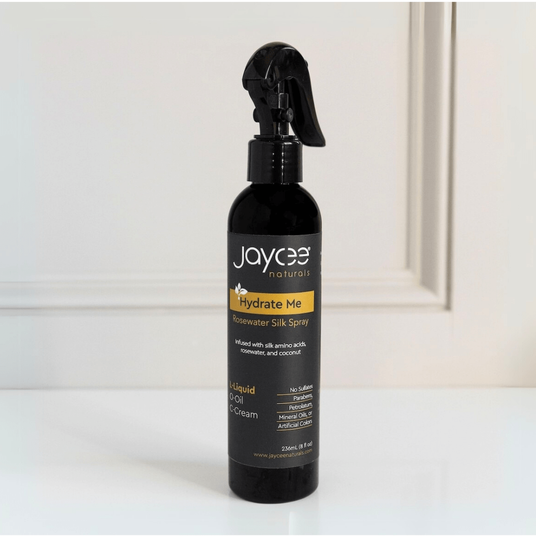 Jaycee Naturals Hydrate Me Rosewater Silk Spray, infused with silk amino acids and free of sulfates, parabens, petrolatum, mineral oils, and artificial colors. 236ml bottle standing on a white countertop.