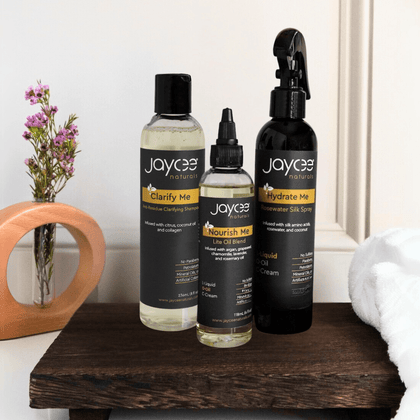 Hair care set for locs, including Nourish Me Hair Oil, Hydrate Me Leave-In Conditioner, and Clarify Me Clarifying Shampoo. One bottle of each product, standing together in a group on a white countertop.