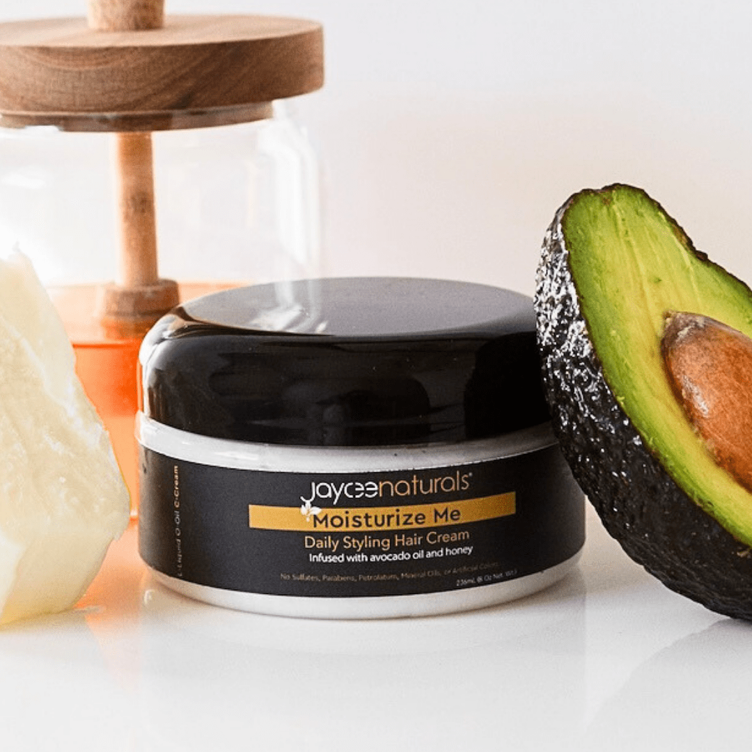 Jaycee Naturals Moisturize Me Hair Cream, infused with avocado oil and honey, and free of sulfates, parabens, petrolatum, mineral oils, and artificial colors. 236ml jar standing on a white countertop surrounded by honey, avocado, and shea butter.