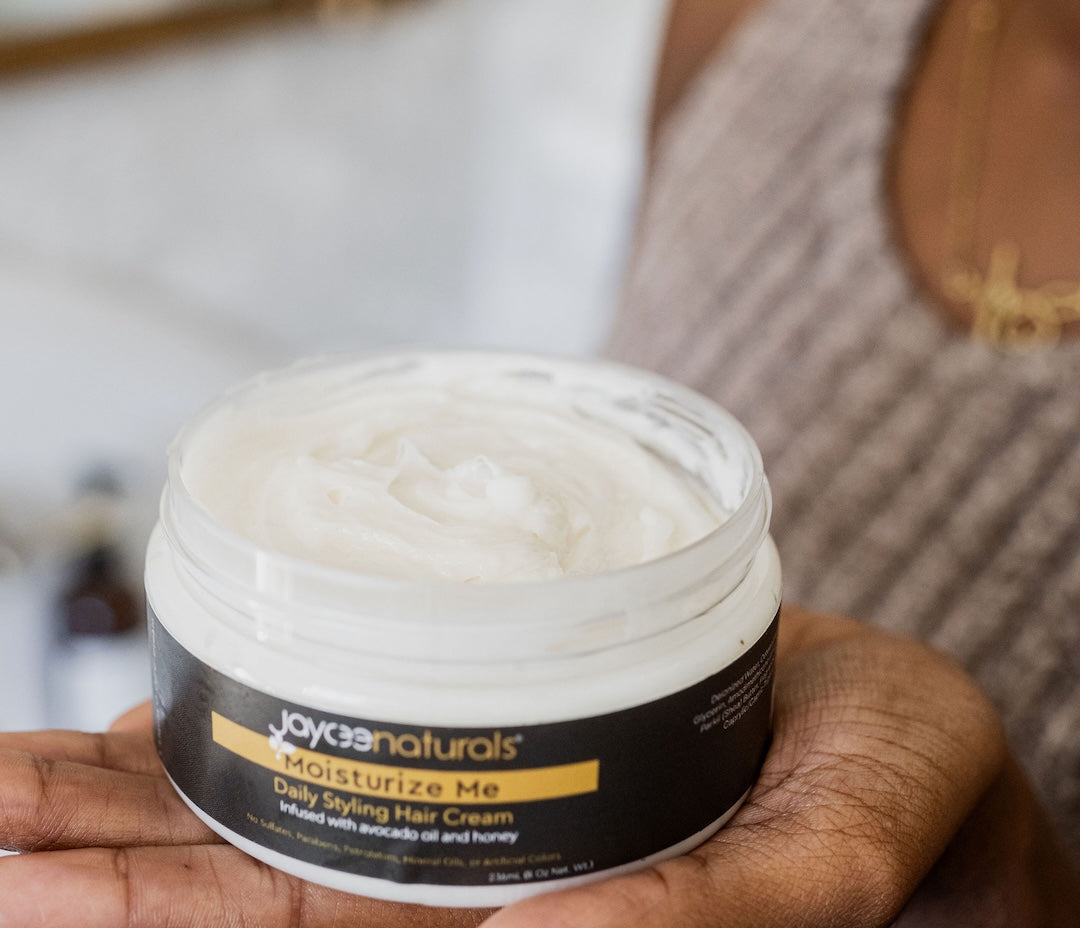 Jaycee Naturals Moisturize Me Hair Cream, infused with avocado oil and honey, and free of sulfates, parabens, petrolatum, mineral oils, and artificial colors. 236ml jar is open and resting in the palm of a hand.