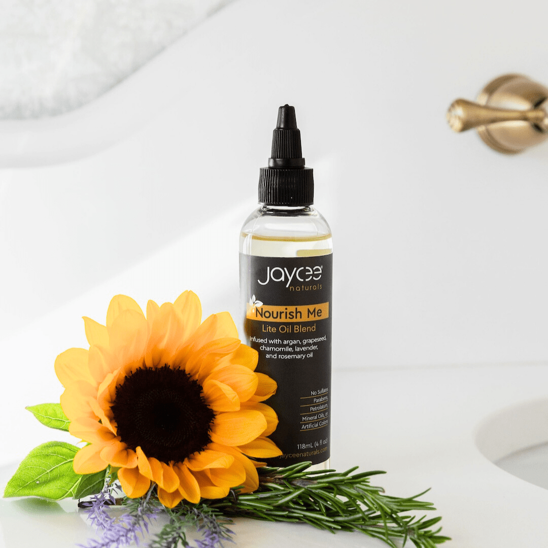 Jaycee Naturals Nourish Me Hair Oil, infused with argan and lavender oils, and free of sulfates, parabens, petrolatum, mineral oils, and artificial colors. 118ml bottle standing on a white countertop surrounded by a sunflower.