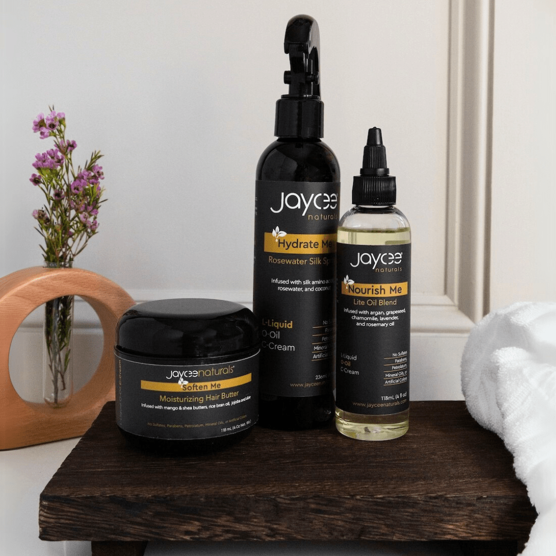 Hair care set for protective styling, including Nourish Me Hair Oil, Hydrate Me Rosewater Silk Spray, and Soften Me Hair Butter. One bottle or jar of each product, standing together in a group on a white countertop.