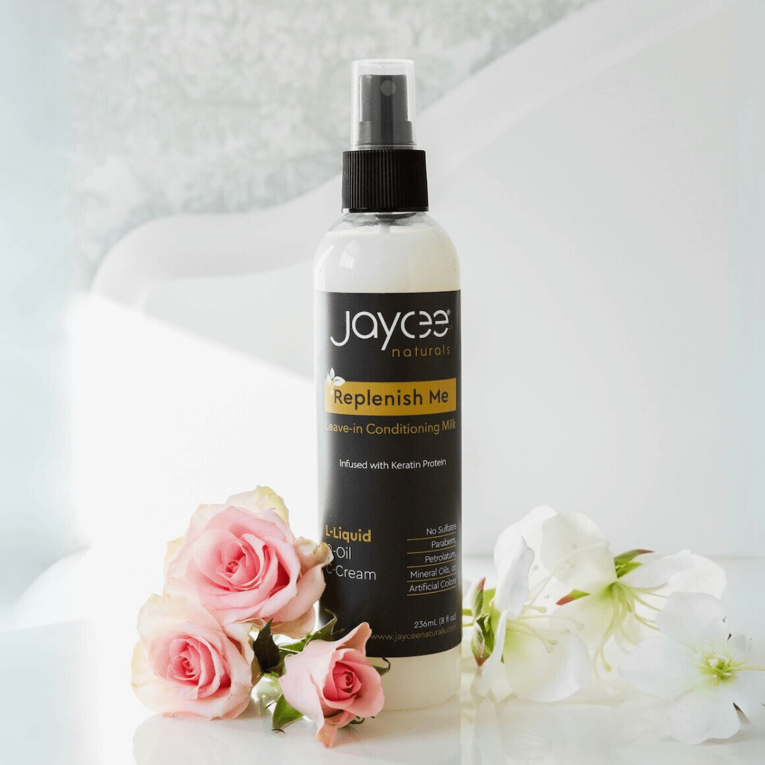 Jaycee Naturals Replenish Me Leave-In Conditioning Milk, infused with keratin protein and free of sulfates, parabens, petrolatum, mineral oils, and artificial colors. 236ml bottle standing on a white countertop and surrounded by white and pink roses.