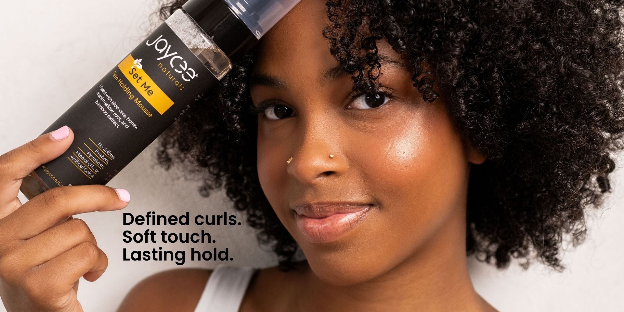 A woman with defined curly hair holds a bottle of Set Me hair mousse. The bottle is black with yellow and white text.