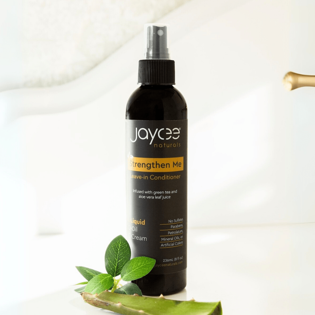 Jaycee Naturals Strengthen Me Leave-In Conditioner, infused with aloe vera leaf juice and free of sulfates, parabens, petrolatum, mineral oils, and artificial colors. 236ml bottle standing on a white countertop surrounded by aloe vera and green tea leaf.